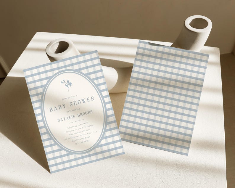 Op de afbeelding: Baby shower uitnodiging met een blauw en wit geruit patroon. De uitnodiging heeft een ovale lijst met bloemendetails en de tekst "Baby Shower" en evenementdetails. Een tweede kaart met hetzelfde patroon is ook zichtbaar.