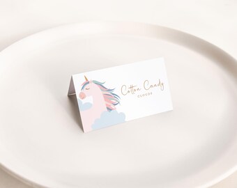 May include: A folded white card with a unicorn illustration and the text "Cotton Candy Clouds" in gold script. The unicorn has a pink body, a gold horn, and a colorful mane. The card is on a white plate.