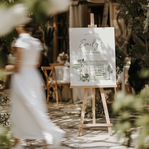 May include: A wooden easel holds a sign with the text "Welcome to Emily's Baby Shower." The sign features an illustration of a bookstore, labelled "Baby's Bookshop." The scene is set outdoors, with a blurred figure in the foreground and greenery.