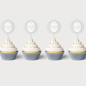 Op de afbeelding: Vier cupcakes met witte glazuur en blauwe papieren vormpjes. Elke cupcake heeft een ronde topper met de tekst "NATALIE'S BABY SHOWER" op een blauw-wit geruite achtergrond. De toppers zijn bevestigd met houten stokjes.