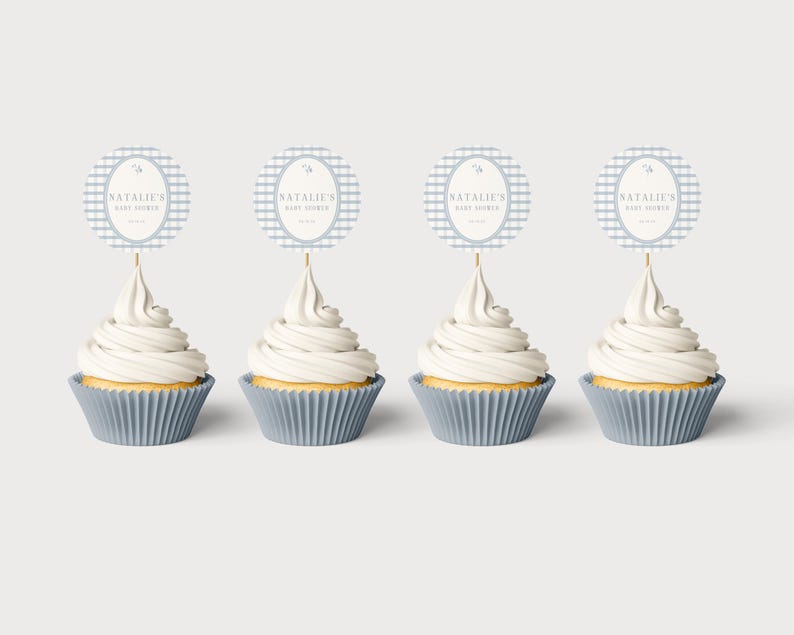 Op de afbeelding: Vier cupcakes met witte glazuur en blauwe papieren vormpjes. Elke cupcake heeft een ronde topper met de tekst "NATALIE'S BABY SHOWER" op een blauw-wit geruite achtergrond. De toppers zijn bevestigd met houten stokjes.
