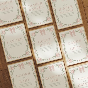 May include: A collection of cream-colored signs with pink text and floral and bow designs. The signs include text such as "Babies are Sweet", "Diaper Raffle", "Favors", and "Guest Book". The signs are arranged on a wooden surface.