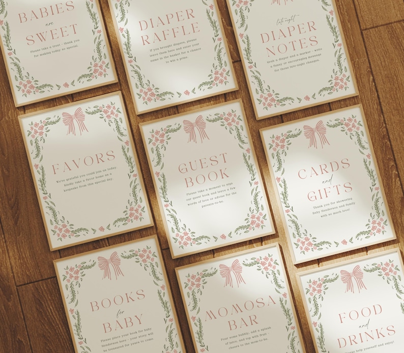 May include: A collection of cream-colored signs with pink text and floral and bow designs. The signs include text such as "Babies are Sweet", "Diaper Raffle", "Favors", and "Guest Book". The signs are arranged on a wooden surface.
