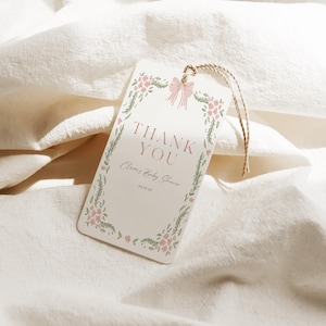 May include: A rectangular cream-colored tag with the words "THANK YOU" in a serif font. The tag is decorated with a floral border and a pink bow. The text "Claire's Baby Shower" and the date are also printed on the tag. A twine string is attached.
