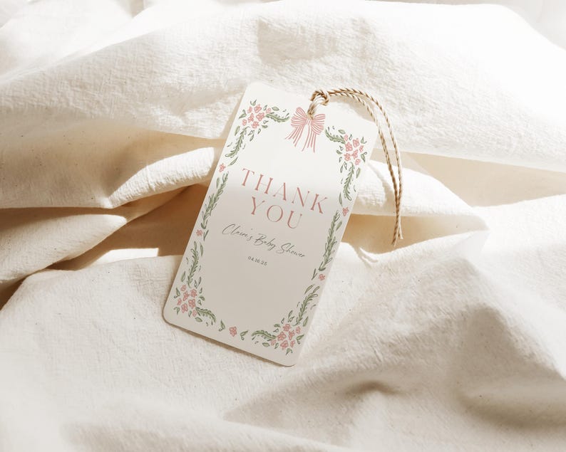 May include: A rectangular cream-colored tag with the words "THANK YOU" in a serif font. The tag is decorated with a floral border and a pink bow. The text "Claire's Baby Shower" and the date are also printed on the tag. A twine string is attached.
