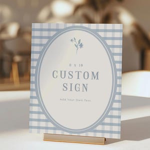 Op de afbeelding: Een aangepast bord met een lichtblauwe gingham patroon achtergrond. Het bord heeft een ovale midden met de woorden "CUSTOM SIGN" in een vet lettertype. Het bord is 20,3 x 25,4 cm en wordt vastgehouden door een goudkleurige metalen standaard.
