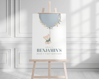 May include: A welcome sign for Benjamin's first birthday, featuring a light blue balloon with floral accents and a teddy bear illustration. The sign is displayed on a wooden easel.