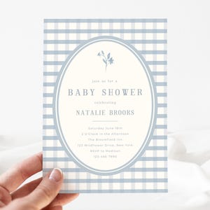 Op de afbeelding: Een uitnodiging voor een babyshower met een lichtblauwe en witte ruitjesachtergrond. De uitnodiging heeft een ovale lijst met de tekst "Baby Shower" en de naam "Natalie Brooks". De uitnodiging wordt door een hand vastgehouden.