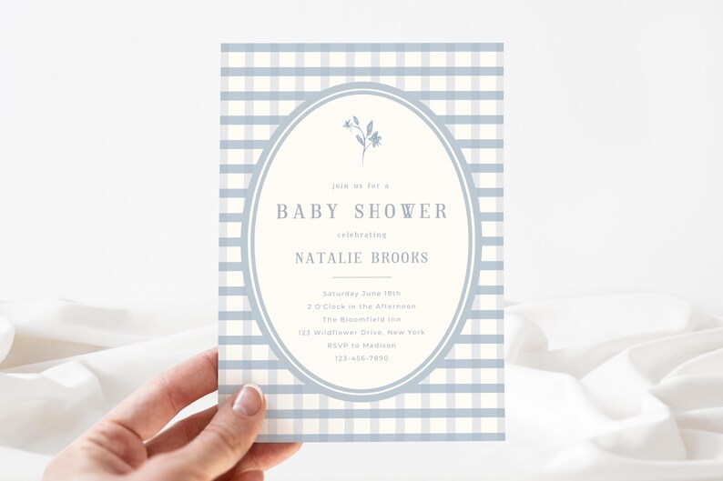 Op de afbeelding: Een uitnodiging voor een babyshower met een lichtblauwe en witte ruitjesachtergrond. De uitnodiging heeft een ovale lijst met de tekst "Baby Shower" en de naam "Natalie Brooks". De uitnodiging wordt door een hand vastgehouden.