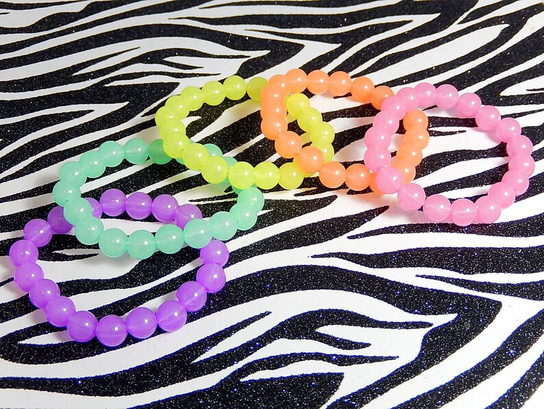 May include: Five colorful bracelets made of round beads. The bracelets are in shades of purple, green, yellow, orange, and pink. The bracelets are arranged in a circle on a black and white zebra print background.