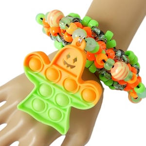 May include: A beaded bracelet featuring a mix of neon green, bright orange, and translucent beads. A silicone ghost-shaped fidget toy charm in orange and lime green with a carved jack-o'-lantern face hangs from the center of the elastic band.