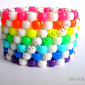 Puede incluir: Un brazalete de cuentas de colores arcoíris con cuentas blancas intercaladas. El brazalete está hecho de cuentas pequeñas y redondas y está enhebrado en un alambre plateado.