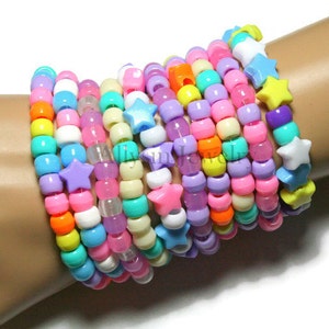 May include: A stack of colourful beaded bracelets with star-shaped charms. The bracelets are made of small, round beads in shades of pink, purple, blue, yellow, and orange. The star charms are in various colours, including white, blue, yellow, and purple.