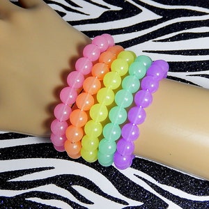May include: A set of six rainbow colored bracelets made of round beads. The bracelets are stacked on a person's wrist.