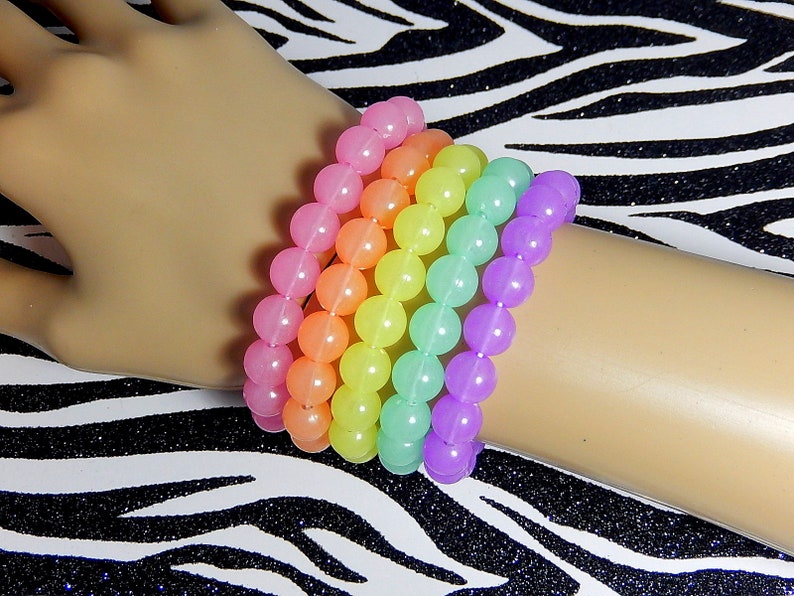 May include: A set of six rainbow colored bracelets made of round beads. The bracelets are stacked on a person's wrist.