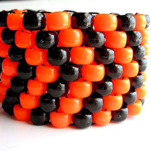 Puede incluir: Un brazalete de cuentas negras y naranjas. El brazalete está hecho de pequeñas cuentas redondas que están enhebradas en un hilo negro. Las cuentas están dispuestas en un patrón de tablero de ajedrez.