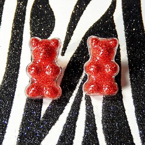 Op de afbeelding: Een paar rode glitter gummy bear oorbellen. De oorbellen zijn op een zwart-witte zebraprint achtergrond.