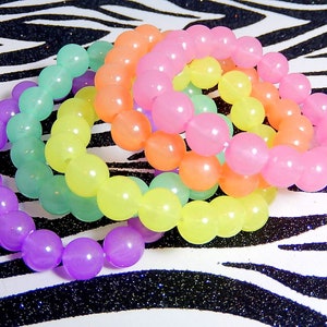 May include: Four colorful bracelets made of round, translucent beads. The bracelets are stacked on top of each other, with the purple bracelet on the bottom, followed by the green, yellow, and pink bracelets.