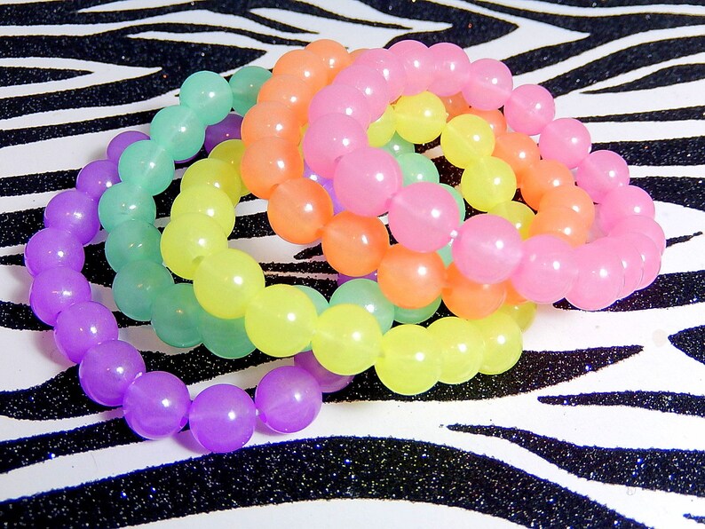 May include: Four colorful bracelets made of round, translucent beads. The bracelets are stacked on top of each other, with the purple bracelet on the bottom, followed by the green, yellow, and pink bracelets.