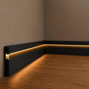 May include: A matte black baseboard molding installed along the corner of a room, featuring a horizontal recessed channel that emits a warm, glowing LED light strip, casting a soft illumination onto the wooden floor surface below the trim.