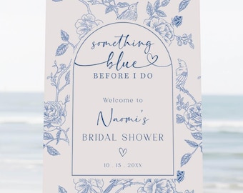 May include: A white sign on a wooden easel with blue floral designs and text that reads "something blue BEFORE I DO Welcome to Naomi's BRIDAL SHOWER ♡ 10.15.20XX". A vase of flowers sits in front of the easel on a sandy beach.