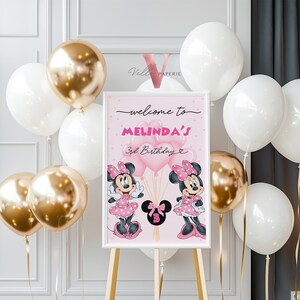 May include: A pink framed sign for a 3rd birthday, featuring Minnie Mouse illustrations and the text "welcome to MELINDA'S 3rd Birthday". The scene is decorated with gold and white balloons.