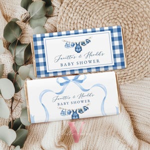 May include: Two baby shower candy bar wrappers. One has a blue gingham pattern, the other has a blue ribbon design. Both wrappers feature the text "Janette's & Harold's BABY SHOWER" and watercolor illustrations of baby clothes.