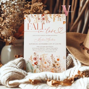 May include: Fall in love bridal shower invitation with a watercolor floral design featuring pumpkins, leaves, and flowers in shades of orange, pink, and green. The invitation text reads "Fall in love" with a heart symbol and includes the bride's name, date, time, and location.