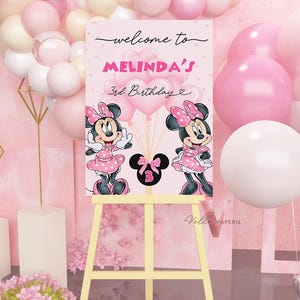 May include: A pink birthday welcome sign on a yellow easel, featuring Minnie Mouse illustrations and the text "welcome to MELINDA'S 3rd Birthday". Pink and white balloons decorate the background.
