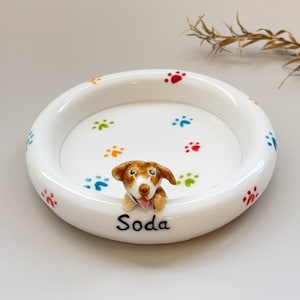 K&ouml;nnte beinhalten: Eine wei&szlig;e Keramik-Haustier-Sch&uuml;ssel mit bunten Pfotenabdr&uuml;cken und einer kleinen, braun-wei&szlig;en Hundefigur. Auf der Sch&uuml;ssel steht der Name "Soda". Ein dekorativer Artikel f&uuml;r Tierbesitzer.