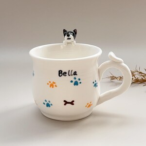 Puede incluir: Taza de cerámica blanca con una figura de perro en la parte superior y un asa curva. La taza está decorada con huellas de patas y un hueso, con el nombre "Bella" escrito en el lateral.