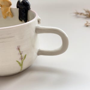 May include: A white ceramic mug with a large handle and a lavender floral design. The rim is painted pink, and two small dog figurines are perched on the edge. The mug is handmade and has a rustic, artisanal feel.