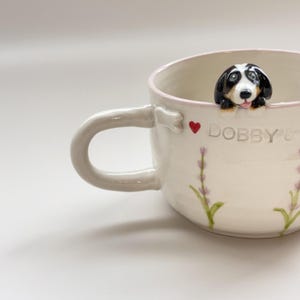 May include: White ceramic mug with a dog figurine on the rim. The mug has a curved handle and is decorated with lavender sprigs and the text "DOBBY &" with a red heart. The dog figurine is black, white, and brown.