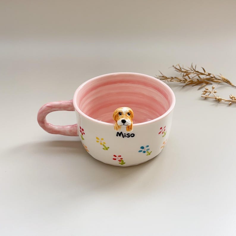 Puede incluir: Una taza de cer&aacute;mica blanca con interior y asa rosas, adornada con dise&ntilde;os florales coloridos. Una peque&ntilde;a y detallada figura de perro est&aacute; adherida al borde. El nombre "Miso" est&aacute; escrito en la taza.