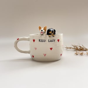 Op de afbeelding: Witte keramische mok met een handvat, versierd met rode en roze harten. De mok heeft twee kleine hondenbeeldjes op de rand en de tekst "RICO LUCY". Een charmant en uniek ontwerp voor hondenliefhebbers.