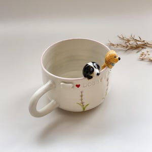 May include: A white ceramic mug with a light pink rim and a curved handle. The mug features two small dog figurines peeking over the rim. The word "DOBBY" is written on the side, along with a red heart and lavender sprigs.