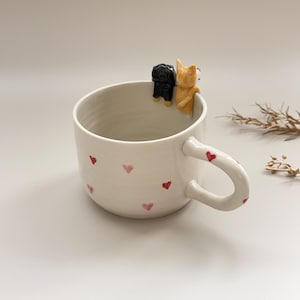 Op de afbeelding: Witte keramische mok versierd met kleine rode hartjes. De handgreep heeft ook rode hartjes. Twee kleine hondenbeeldjes, een zwarte en een bruine, zitten op de rand. Een unieke koffiemok.