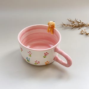 Puede incluir: Una taza de cer&aacute;mica blanca con interior y asa rosas, adornada con dise&ntilde;os florales coloridos. Una peque&ntilde;a figura naranja se encuentra en el borde. La taza es ideal para caf&eacute;, t&eacute; u otras bebidas.