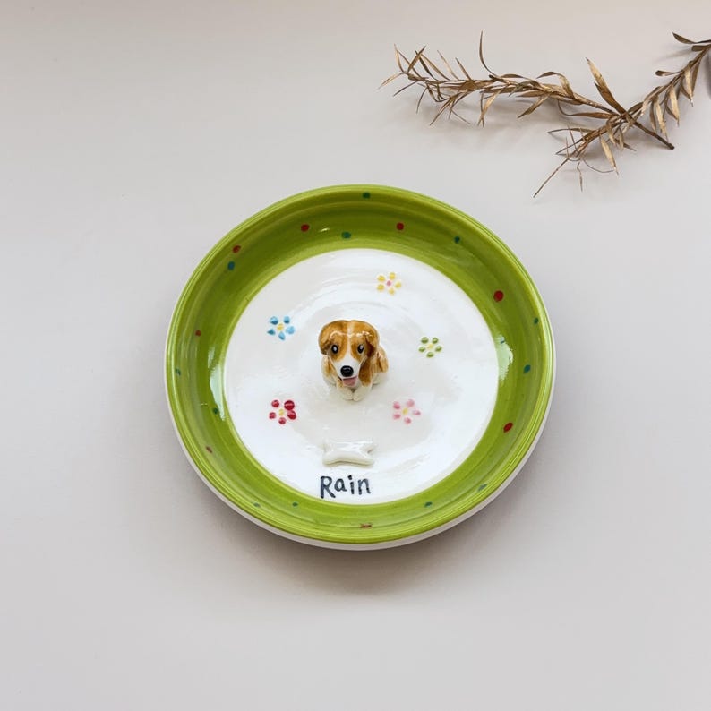 May include: A ceramic dog bowl with a green rim and a white interior. The bowl features a 3D dog figurine in the center, surrounded by colorful paw prints and flower designs. The name "Rain" is written in blue script.
