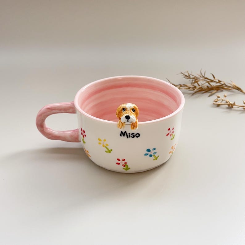 Puede incluir: Una taza de cer&aacute;mica blanca con interior y asa rosas, decorada con dise&ntilde;os florales coloridos. Una peque&ntilde;a y detallada figura de perro est&aacute; posada en el borde, con el nombre "Miso" debajo. Una pieza encantadora y &uacute;nica.
