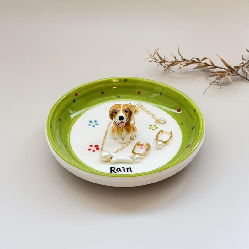 May include: A ceramic trinket dish with a green rim and a white interior. The dish features a small, detailed dog figurine and paw prints. A necklace, earrings, and a ring are displayed on the dish. The word "Rain" is written on the dish.
