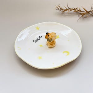 Peut inclure: Une assiette en c&eacute;ramique blanche aux bords irr&eacute;guliers, orn&eacute;e d'&eacute;toiles jaunes et d'un croissant de lune. Une petite figurine d&eacute;taill&eacute;e de chien est au centre. Le nom "Oakley" est &eacute;crit en script noir sur l'assiette.