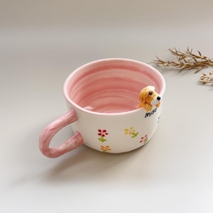 Puede incluir: Una taza de cer&aacute;mica blanca con interior y asa rosas. La taza presenta una peque&ntilde;a figura tridimensional de un perro golden retriever en el borde. Tambi&eacute;n hay dise&ntilde;os florales y la palabra "Miso" en la taza.