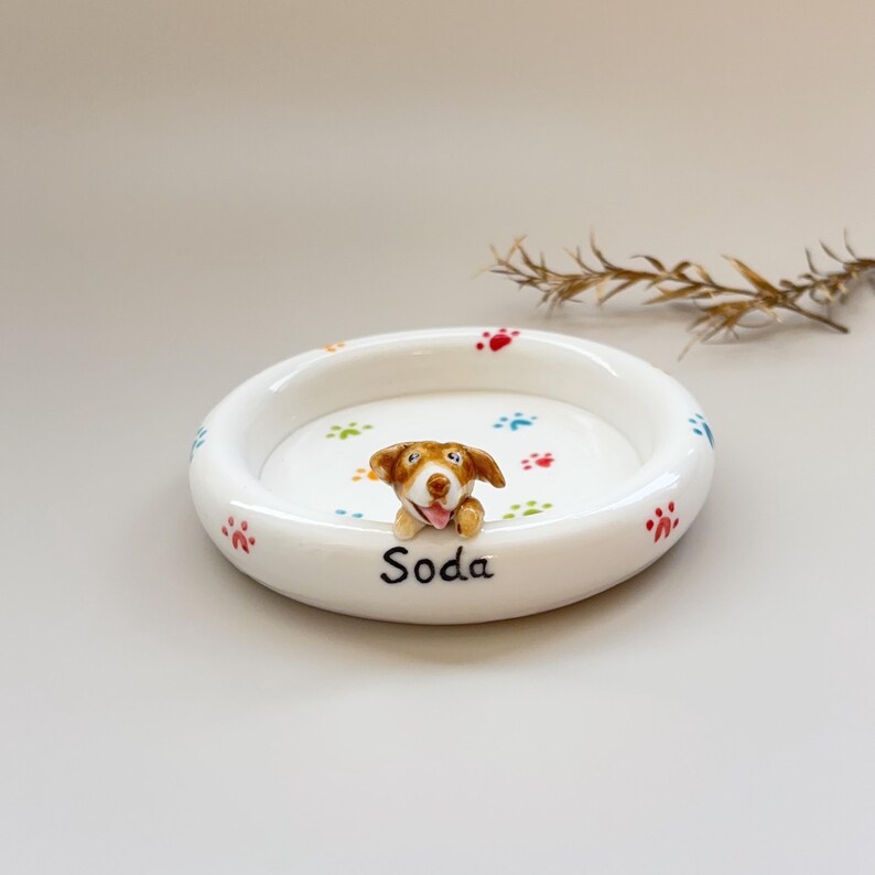 K&ouml;nnte beinhalten: Eine wei&szlig;e Keramik-Haustier-Sch&uuml;ssel mit erh&ouml;htem Rand, verziert mit bunten Pfotenabdr&uuml;cken und einer kleinen, l&auml;chelnden Hundefigur. Die Sch&uuml;ssel ist mit dem Namen "Soda" in schwarzer Schrift personalisiert.
