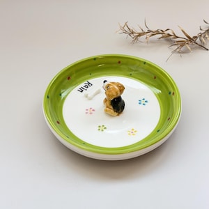 May include: A ceramic dog bowl with a green rim and a white interior. The bowl features a small, decorative dog figurine and the word "Rain". Colorful dots and flower designs are scattered around the bowl.