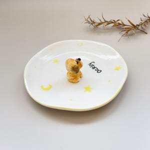 Peut inclure: Une petite assiette en c&eacute;ramique blanche avec un bord jaune et des motifs d'&eacute;toiles et de croissants de lune. Une petite figurine de chien de couleur beige est au centre. Le mot "Oakley" est &eacute;crit en noir sur l'assiette.