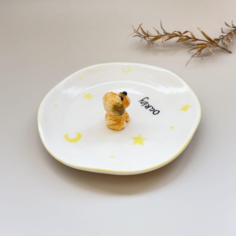 Peut inclure: Une petite assiette en c&eacute;ramique blanche avec un bord jaune et des motifs d'&eacute;toiles et de croissants de lune. Une petite figurine de chien de couleur beige est au centre. Le mot "Oakley" est &eacute;crit en noir sur l'assiette.