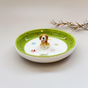 May include: A ceramic dog-themed dish with a white interior and a green rim. The dish features a small, detailed figurine of a dog and a bone-shaped accent. Colorful dots and flower-shaped designs decorate the interior.
