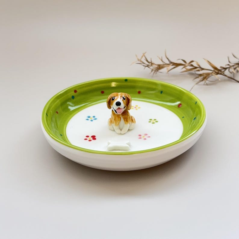 May include: A ceramic dog-themed dish with a white interior and a green rim. The dish features a small, detailed figurine of a dog and a bone-shaped accent. Colorful dots and flower-shaped designs decorate the interior.