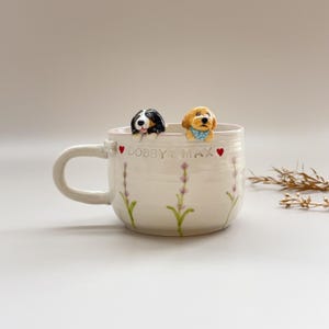 May include: A white ceramic mug with a handle, featuring two dog figurines peeking over the rim. The mug is decorated with lavender sprigs and the text "DOBBY & MAX". The dogs are a black and white Bernese Mountain Dog and a golden Labradoodle.